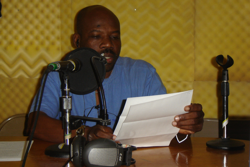 Un homme assis devant un micro de radio, lisant un texte d'une feuille qu'il tient en mains