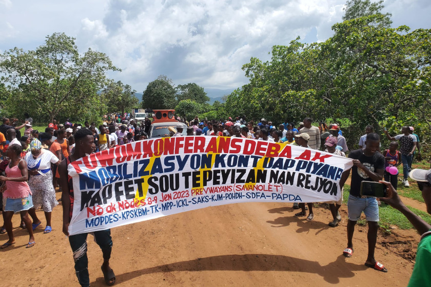 Manifestation d'haïtiens sur une route de terre, en avant plan deux hommes portent une banderole avec un texte en créole haïtien