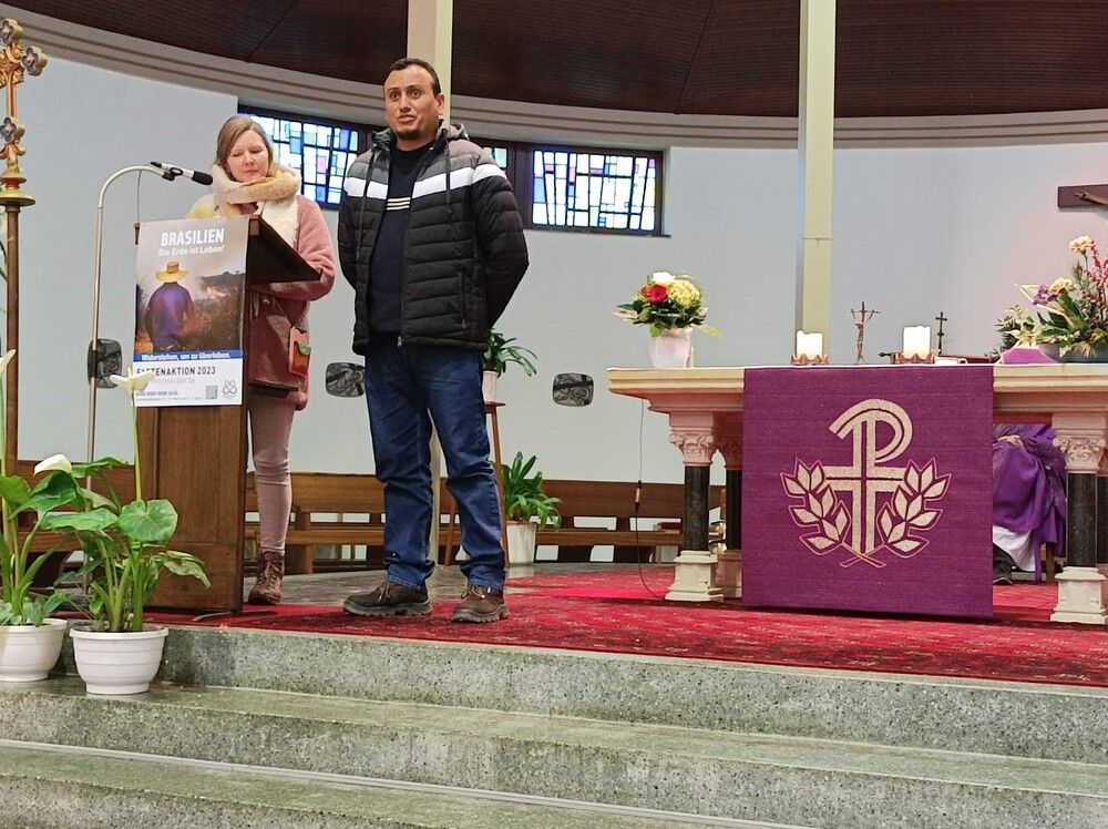 Dans une église, deux personnes devant un microphone. L'affiche de la campagne de Carême 2023 sur le devant du pupitre.