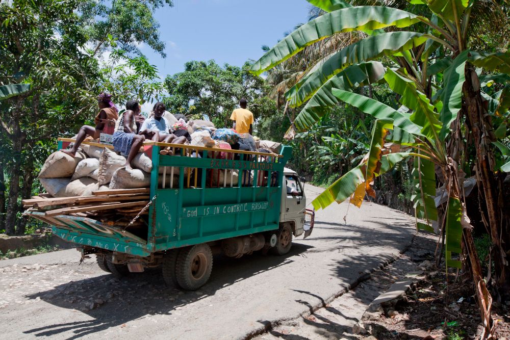 Un camion sur une route entre bananiers, chargé de planches en bois et de sacs en jute, quelques personnes assises au dessus.