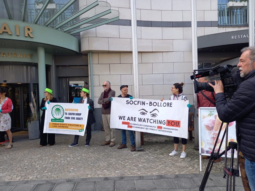 Un groupe de manifestants avec des bannières : SOCFIN-BOLLORE : WE ARE WATCHING YOU