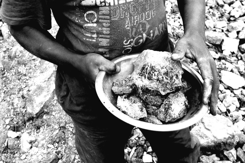 photo noir et blanc : les mains d'un ouvrier tinnent un content en métal rempli de morceaux de roche.