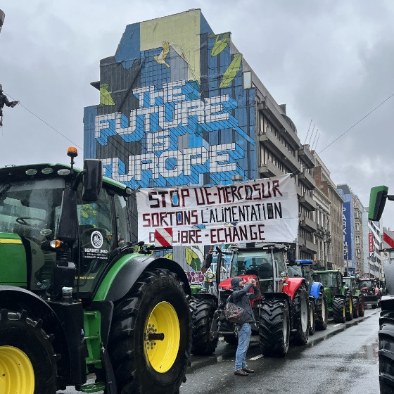 Tracteurs sur un boulevard. En arrière plan une frèsque murale avec le texte "The future is Europe". En avant plan un tracteur avec une banderole : "Stop EU-Mercosur - sortons l'alimentation du libre échange"