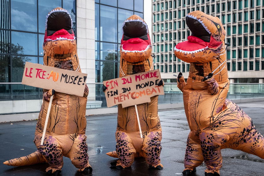 Trois personnes en constume de dinosaures, deux avec des pancartes : "TCE au musée" et "Fin du monde / Fin du monde / même combat"