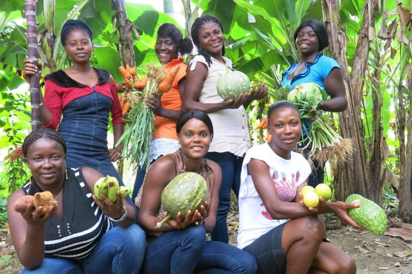 Un groupe de femmes souriantes pose entre des bananiers. Chacune tient un légume ou fruit différent dans les mains.