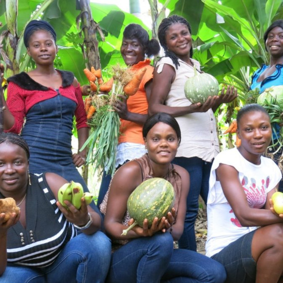 Un groupe de femmes souriantes pose entre des bananiers. Chacune tient un légume ou fruit différent dans les mains.