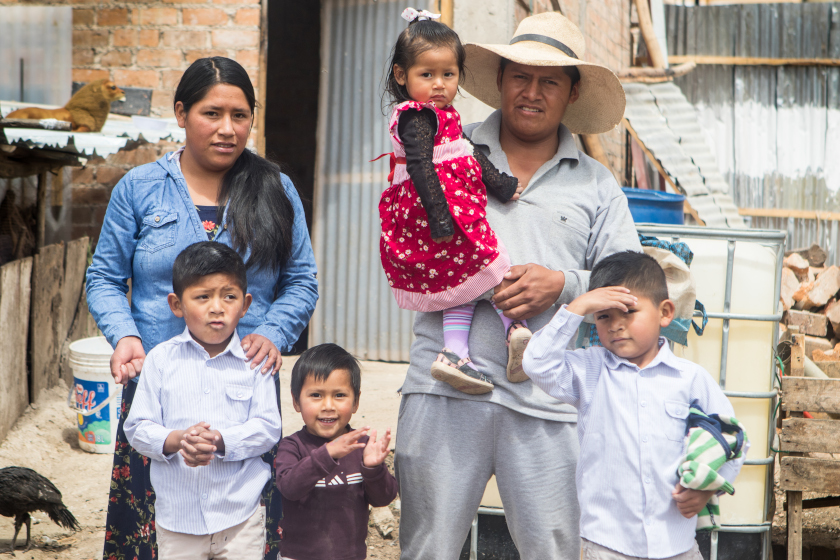 Une famille avec 4 jeunes enfants devant une maison modeste avec une porte en tôle.