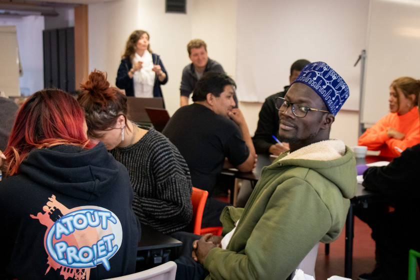 des personnes assises en discussion, deux personnes en arrière plan animent le groupe, en avant plan une personne regarde dans la caméra