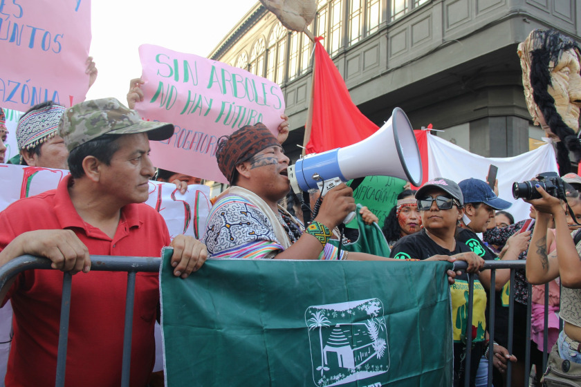 Une manifestatio. Des gens avec des pancartes derrière une barrière nadar. Une personne parle dans un mégaphone.