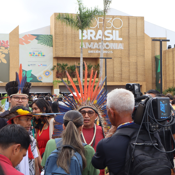 Un caméraman filme une personne en tenue indigène dans une foule devant un bâtiment avec l'enseigne COP 30 Brasil Amazonia.