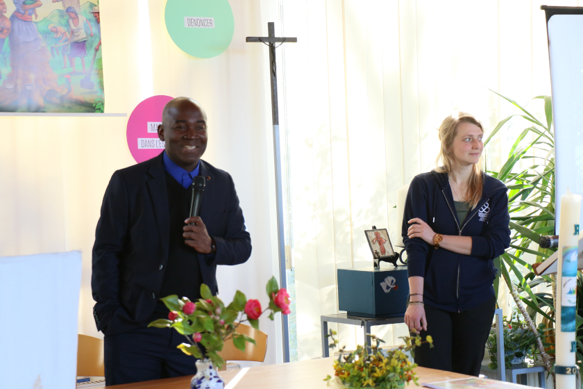Une personne debout parle dans un micro devant un public hors champs. Une deuxième personne à coté porte le logo d'Entraide et Fraternité sur son pull. Sur le mur derrière eux on voit une croix et un bout de l'affiche de la campagnes de Carême