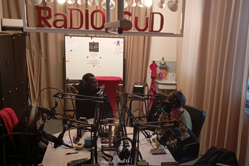 Deux personnes avec des casques devant des micros, assise devant une dable technique dans un studio son. Au mur une enseigne "Radio Sud"