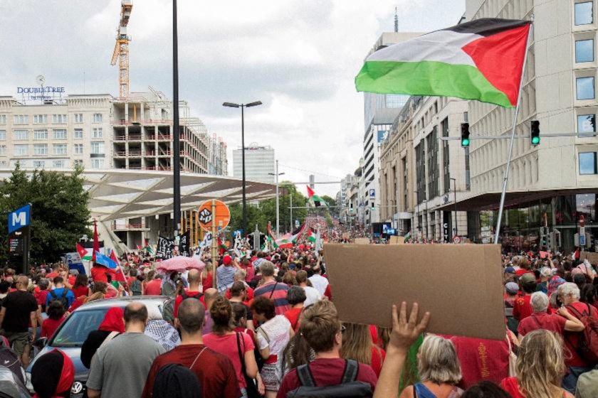 Une grande foule remplit une large avenue urbaine lors d’une manifestation. De nombreuses personnes brandissent des pancartes et des drapeaux, dont plusieurs drapeaux aux couleurs noire, blanche, verte et rouge.