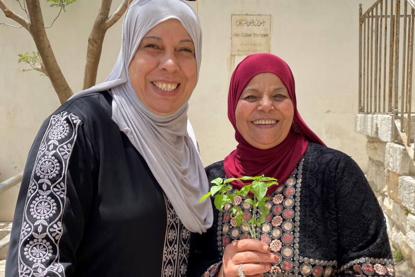 Deux femmes souriantes devant un mur beige ensoleillé. Une tient un brin d'un herbe fraischement cueilli.