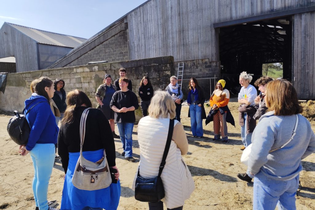 Groupe debout en cercle devant une étable, écoutant une femme au centre du groupe.