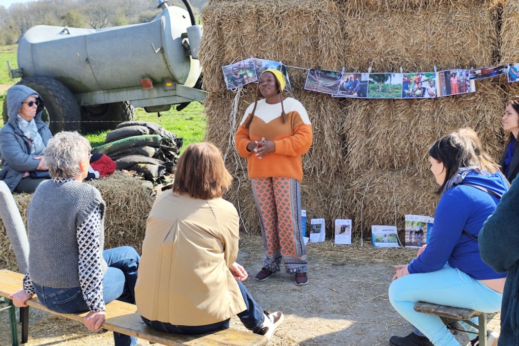 Femme parlant devant un petit groupe assis, avec des photos accrochées sur des bottes de foin. En arrière plan un engin agricole.