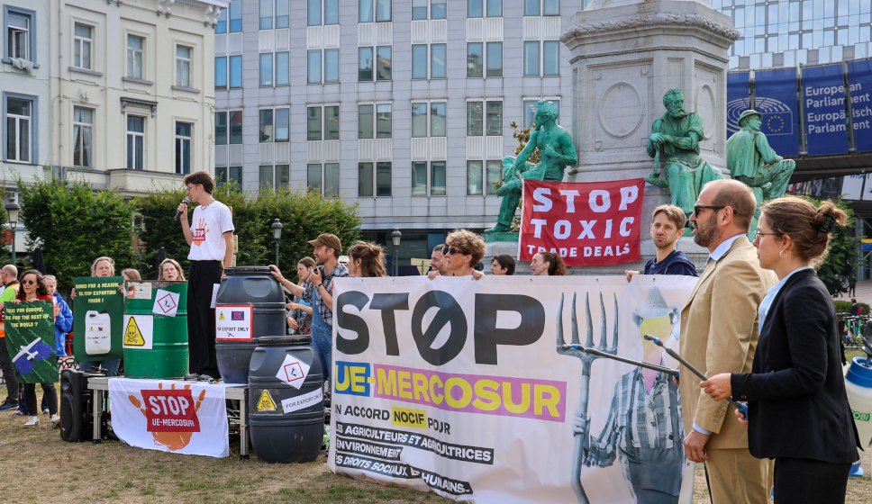 Manifestation devant le Parlement européen avec des pancartes “STOP UE‑MERCOSUR” et “STOP TOXIC TRADE DEALS”, des barils symboliques et un groupe de participants rassemblés.