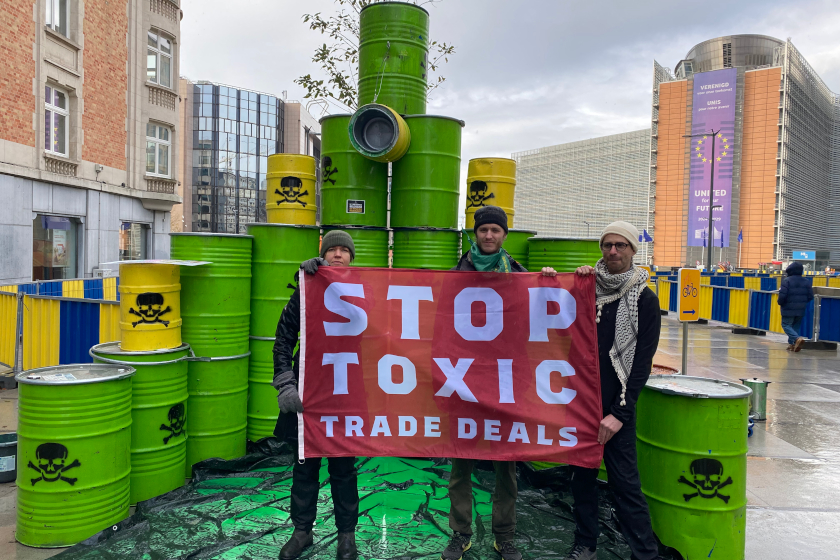 Trois personnes tiennent une banderole “STOP TOXIC TRADE DEALS” devant un assemblage de barils verts décorés de symboles toxiques, devant le bâtiment de la commission européenne.